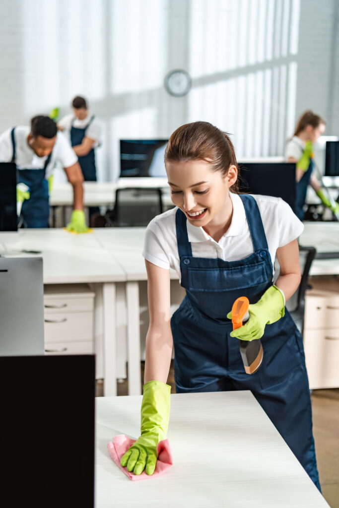 A group of people wearing blue overalls and yellow gloves cleaning desks in an office.