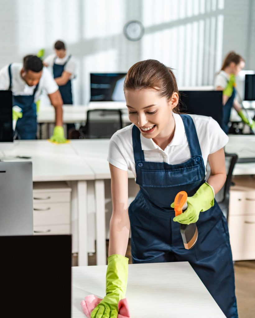 A group of people wearing blue overalls and yellow gloves cleaning desks in an office.